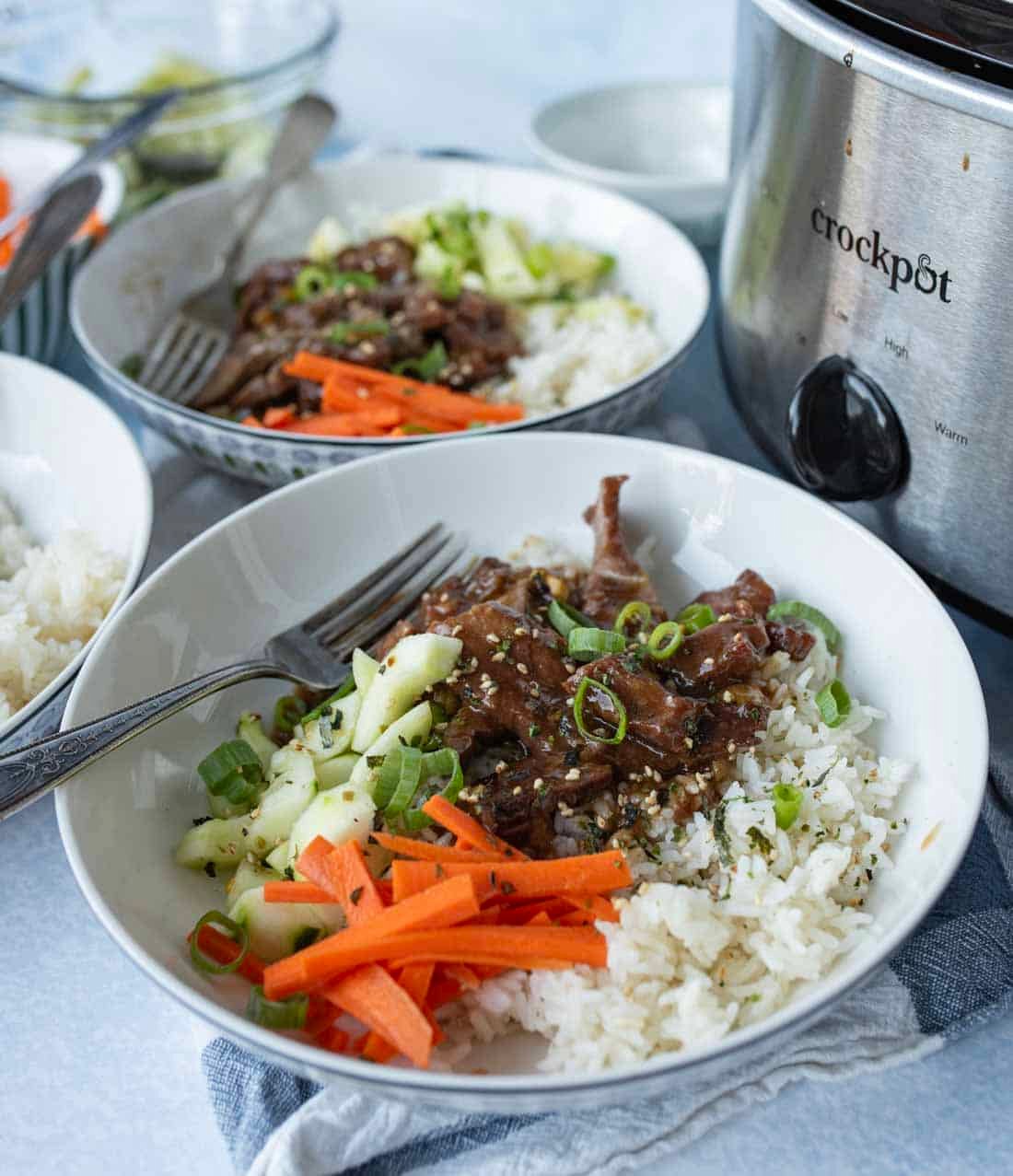 A bowl of white rice topped with shredded beef, sliced green onions, sesame seeds, julienned carrots, and cucumber, with a fork. A crockpot and another bowl of food are in the background.