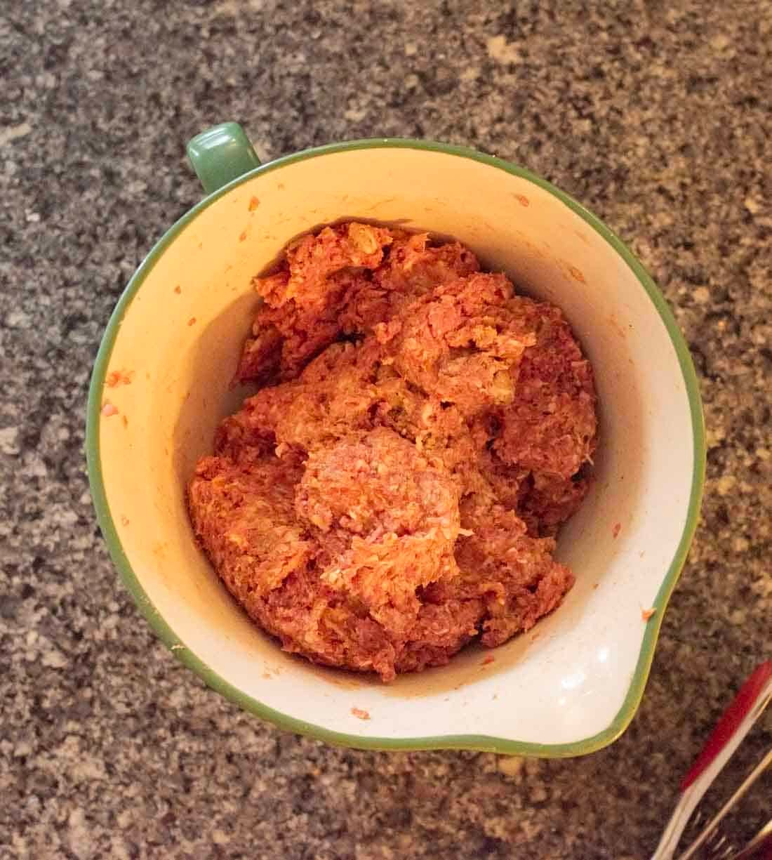 A green mixing bowl filled with seasoned raw ground meat mixture sits on a speckled kitchen countertop.