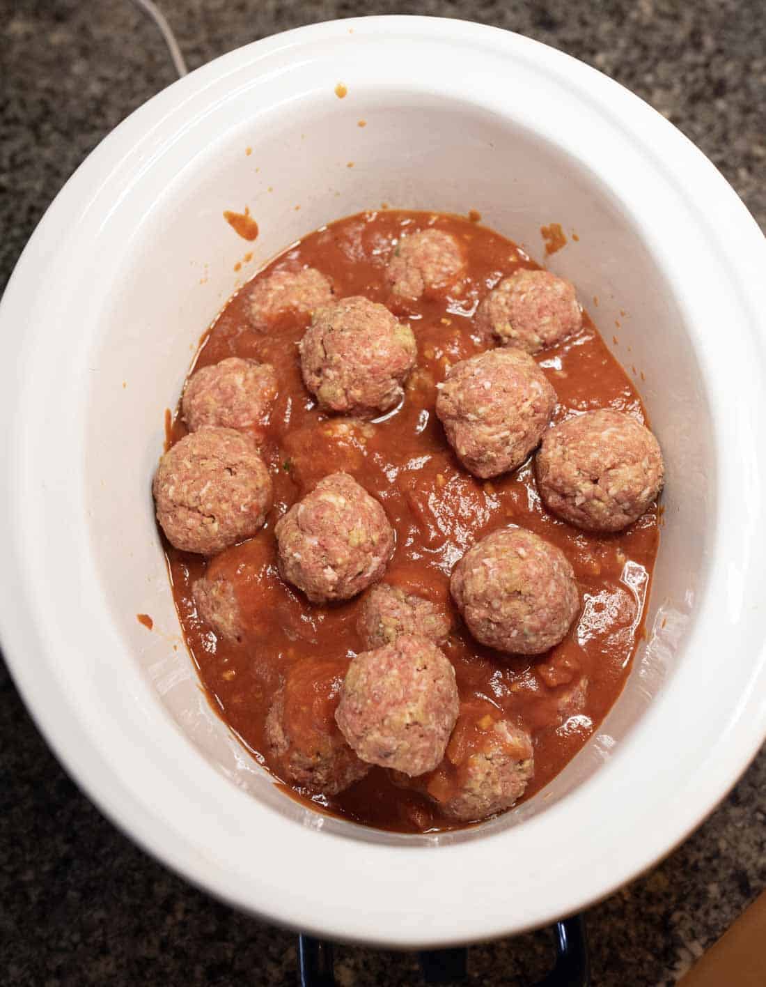 Uncooked meatballs sit in tomato sauce inside a white slow cooker, placed on a granite countertop.