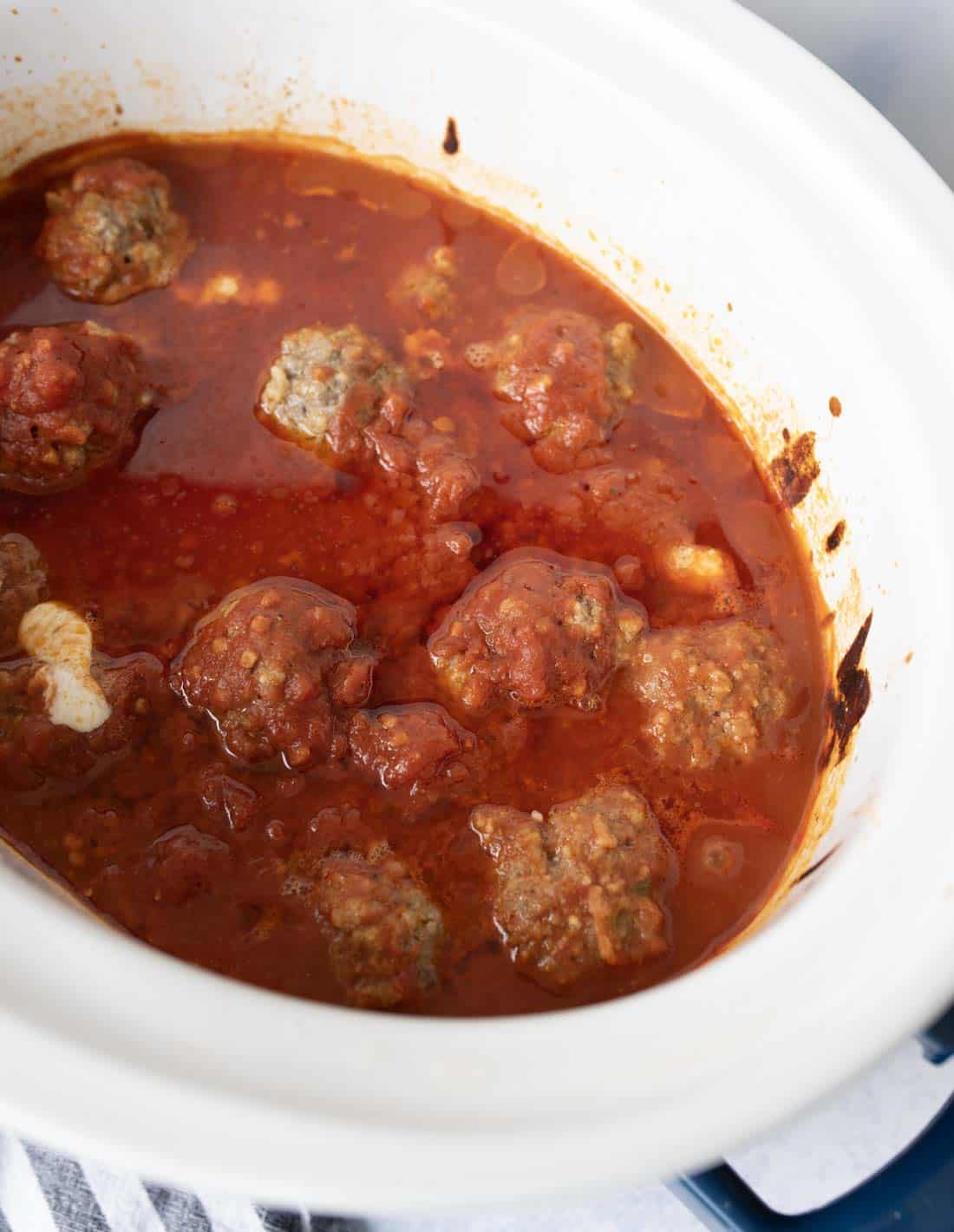 Meatballs in tomato sauce simmering inside a white slow cooker, with some sauce splattered along the sides of the pot.
