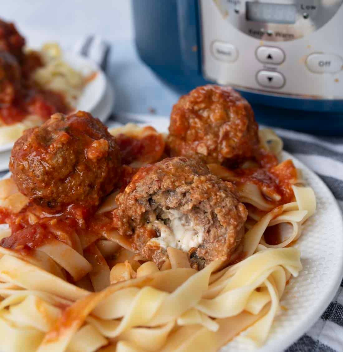 A plate of fettuccine noodles topped with tomato sauce and meatballs, one of which is cut open to show a cheese-filled center. An electric pressure cooker is visible in the background.
