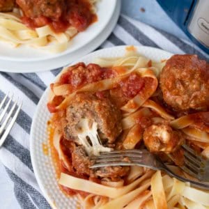 A plate of fettuccine pasta topped with tomato sauce and mozzarella stuffed meatballs, with a fork cutting into a meatball; another plate with the same meal is in the background.