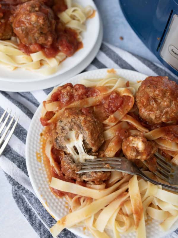 A plate of fettuccine pasta topped with tomato sauce and mozzarella stuffed meatballs, with a fork cutting into a meatball; another plate with the same meal is in the background.