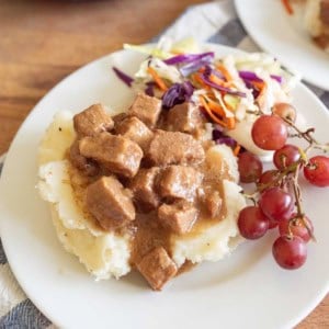 A plate of mashed potatoes topped with slow cooker steak bites, served with coleslaw and a small bunch of red grapes on the side.