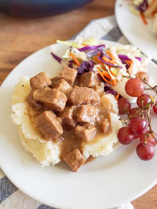 A plate of mashed potatoes topped with slow cooker steak bites, served with coleslaw and a small bunch of red grapes on the side.