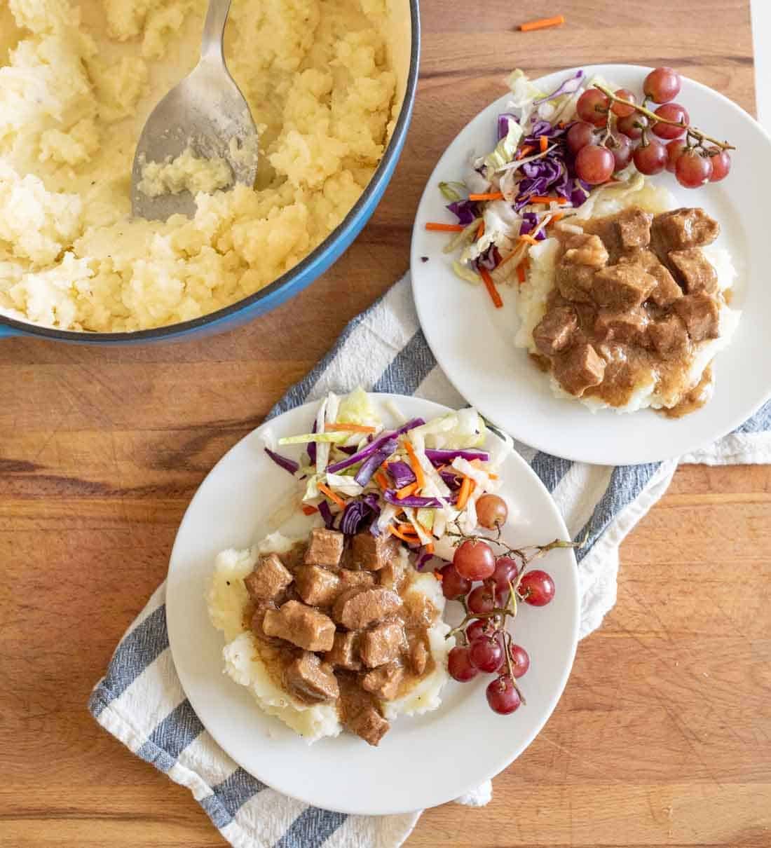Two plates with mashed potatoes topped with brown gravy and meat cubes, served with a grape bunch and colorful coleslaw. A pot of mashed potatoes and a serving spoon sit nearby on a wooden surface with a striped towel.