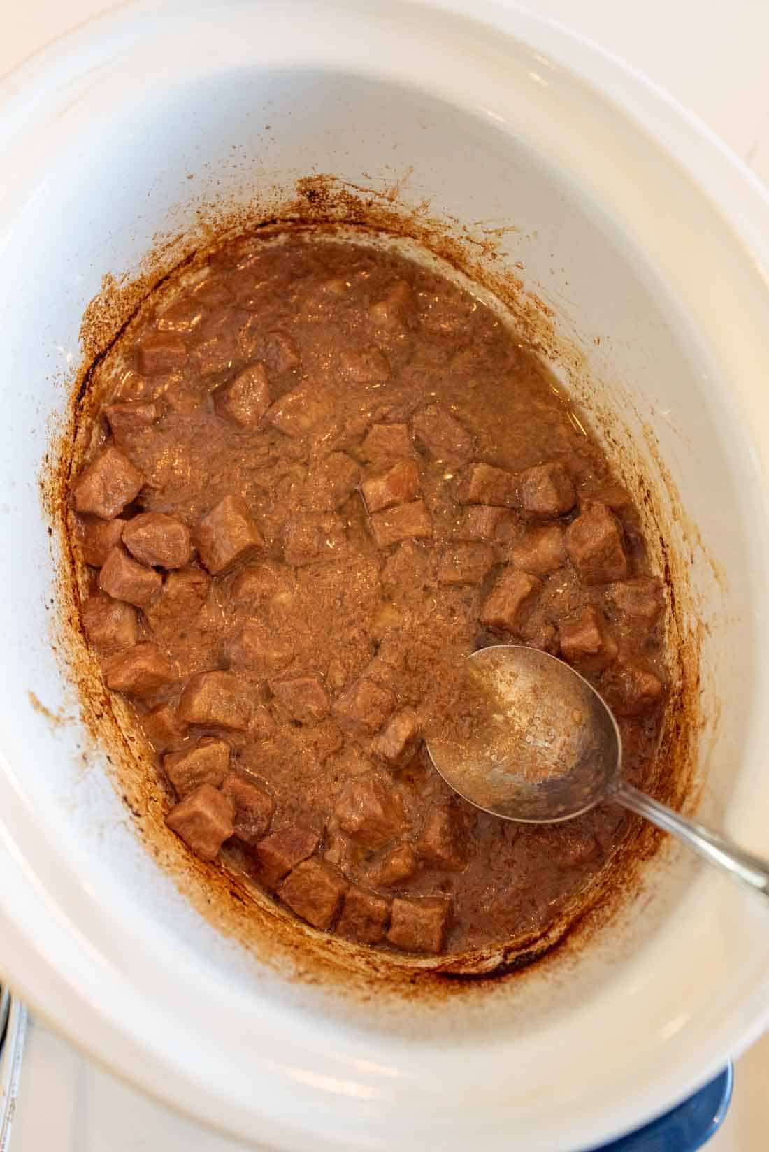 Chunks of beef in brown gravy cooking in a white slow cooker, with a metal spoon resting inside the pot.