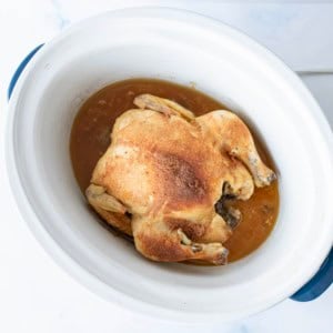 A whole cooked chicken with seasoned skin sits in a white slow cooker, surrounded by brown cooking juices.