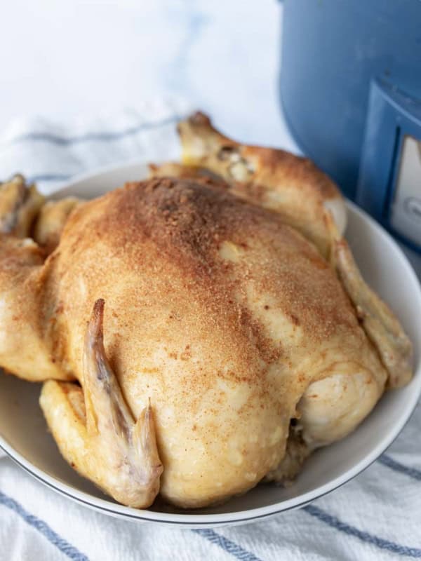 A slow cooker whole chicken seasoned with spices sits in a white bowl on a striped towel, with part of a blue slow cooker visible in the background.