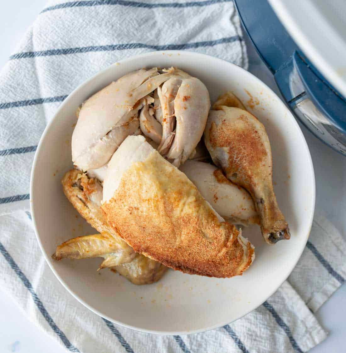A bowl of cooked chicken pieces, including a drumstick, breast, and thigh, sits on a white plate atop a striped kitchen towel, with part of a slow cooker visible in the corner.