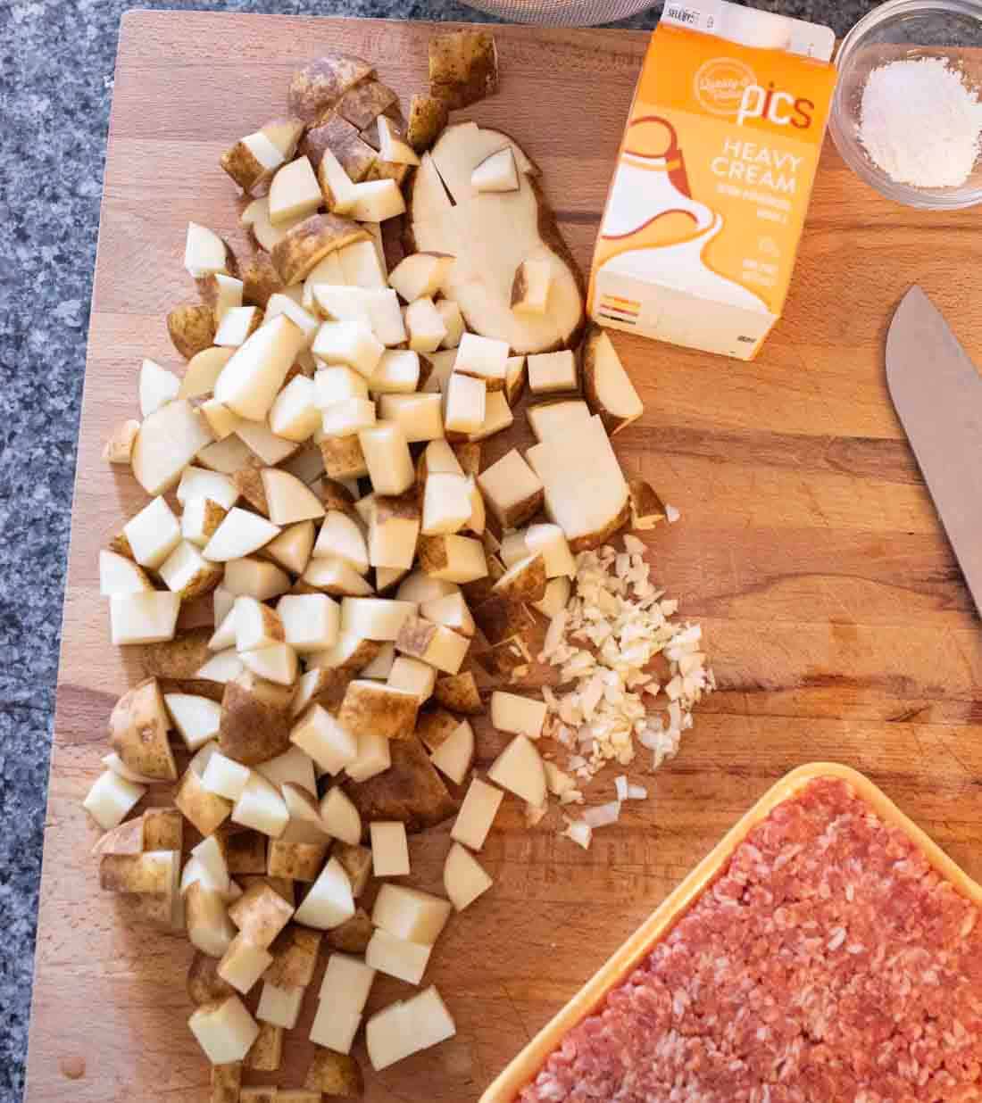 Overhead view of a wooden cutting board with diced potatoes, chopped onions, a knife, a package of ground meat, a small bowl of salt, and a carton of heavy cream on a gray countertop.