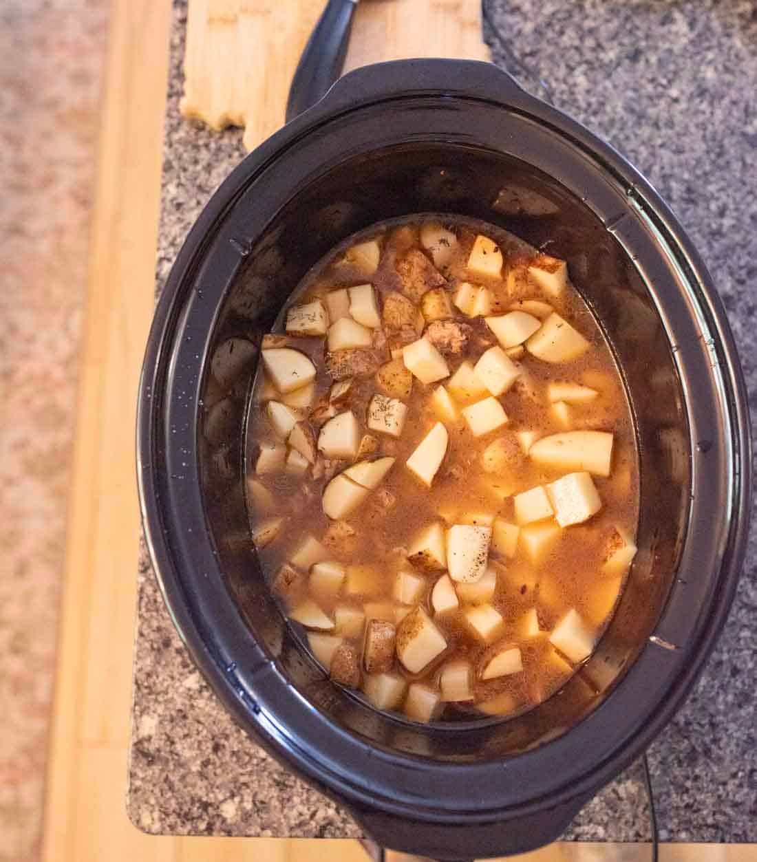 A slow cooker filled with a mixture of diced potatoes, beef chunks, and broth sits on a kitchen countertop next to a wooden cutting board.