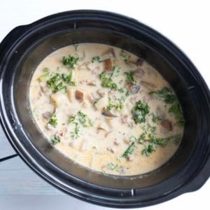 A slow cooker filled with creamy soup containing chopped kale, diced potatoes, sausage, and a light broth, viewed from above.