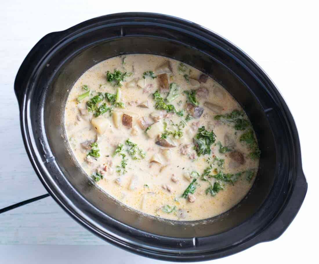 A slow cooker filled with creamy soup containing chopped kale, diced potatoes, sausage, and a light broth, viewed from above.