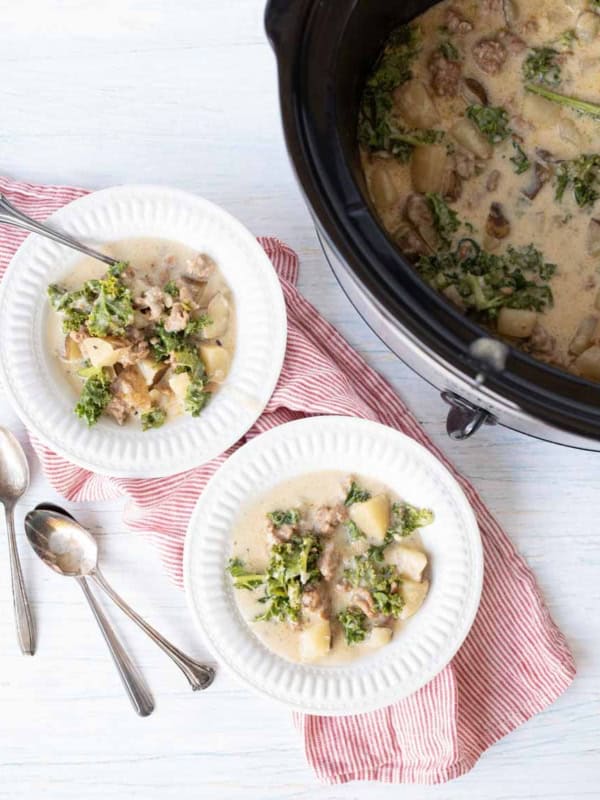 Two white bowls of creamy sausage potato soup with sausage, potatoes, and kale sit on a red-striped cloth next to a slow cooker filled with the same soup. Two spoons lie beside the bowls on a light surface.