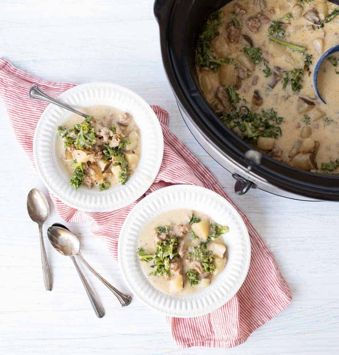 Two white bowls of creamy sausage potato soup with sausage, potatoes, and kale sit on a red-striped cloth next to a slow cooker filled with the same soup. Two spoons lie beside the bowls on a light surface.