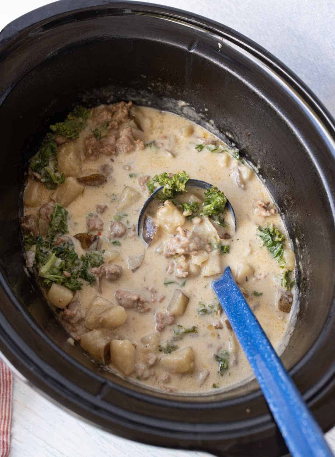 A slow cooker filled with creamy soup containing chunks of potato, ground sausage, and green leafy kale, with a blue ladle resting inside.