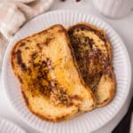 Two slices of golden-brown sourdough French toast with syrup on a white plate, next to a cup of tea, cinnamon sticks, a fork and knife, a small bowl of cinnamon, and a beige napkin.