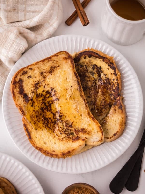 Two slices of golden-brown sourdough French toast with syrup on a white plate, next to a cup of tea, cinnamon sticks, a fork and knife, a small bowl of cinnamon, and a beige napkin.