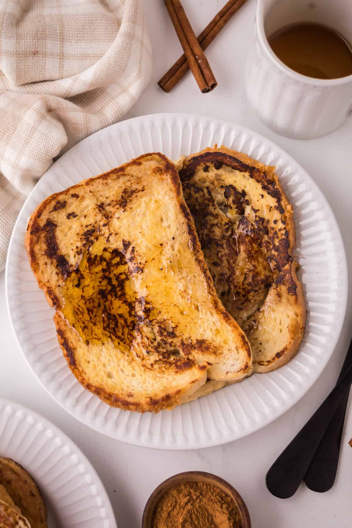 Two slices of golden-brown sourdough French toast with syrup on a white plate, next to a cup of tea, cinnamon sticks, a fork and knife, a small bowl of cinnamon, and a beige napkin.