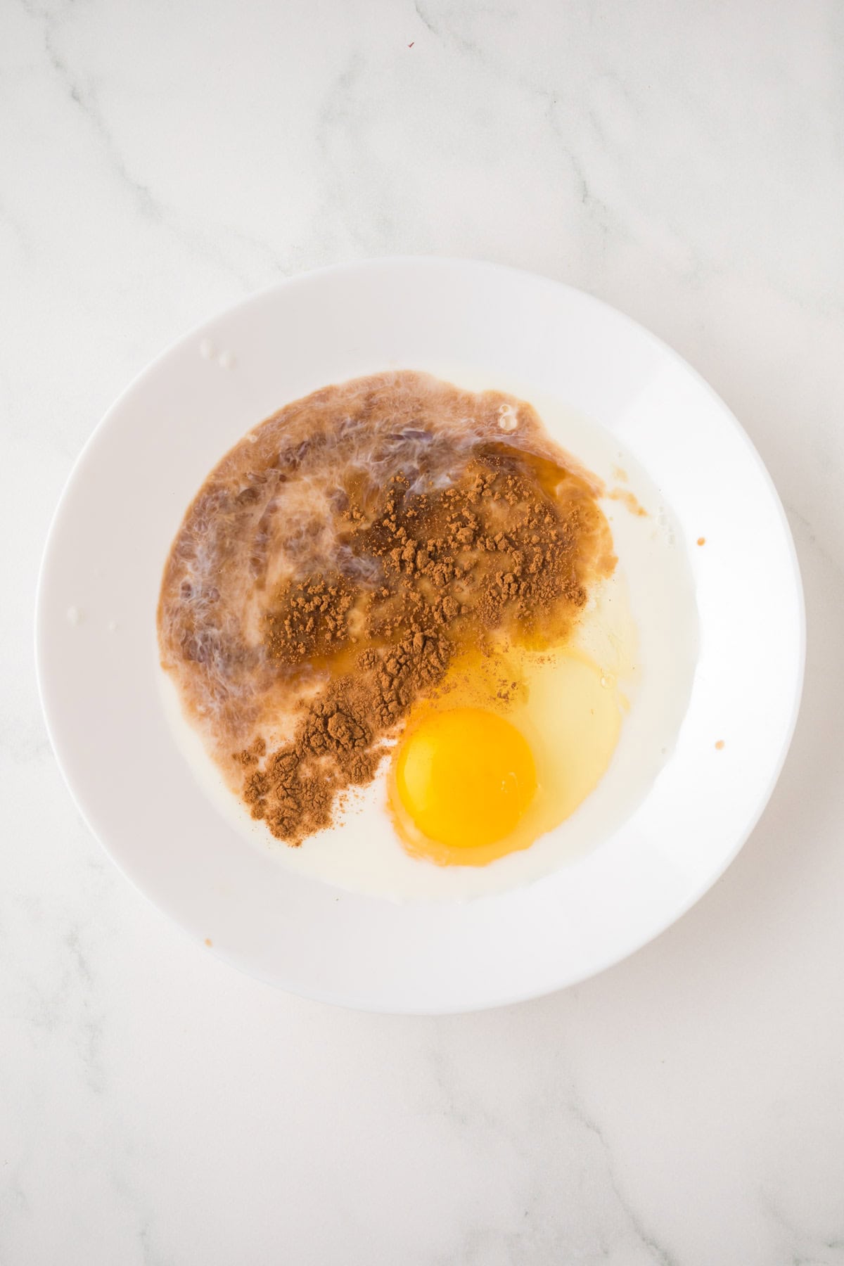 A white plate with an uncooked egg, milk, and cocoa powder sitting separately on a white marble surface, ready to be mixed for decadent sourdough French toast.