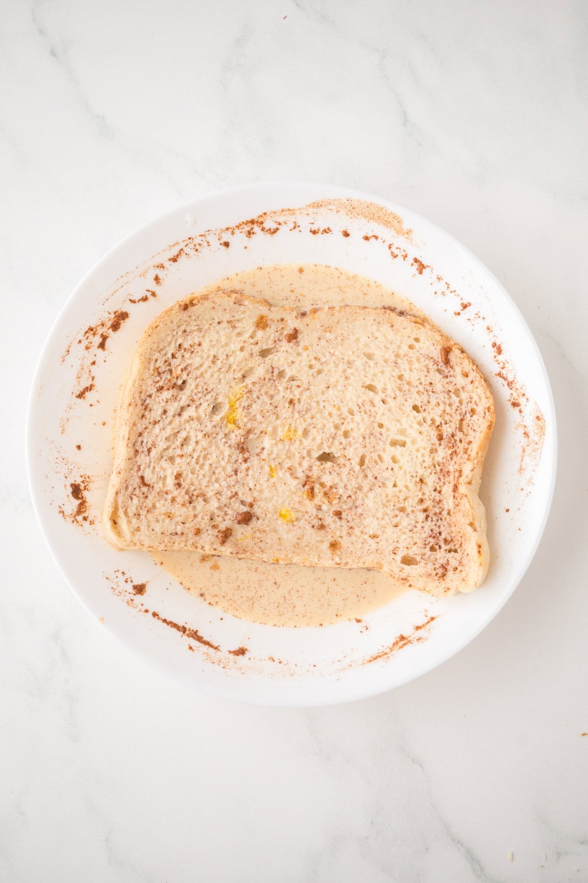 A slice of sourdough French toast soaked in an egg and cinnamon mixture sits on a white plate, which is placed on a white marble surface.