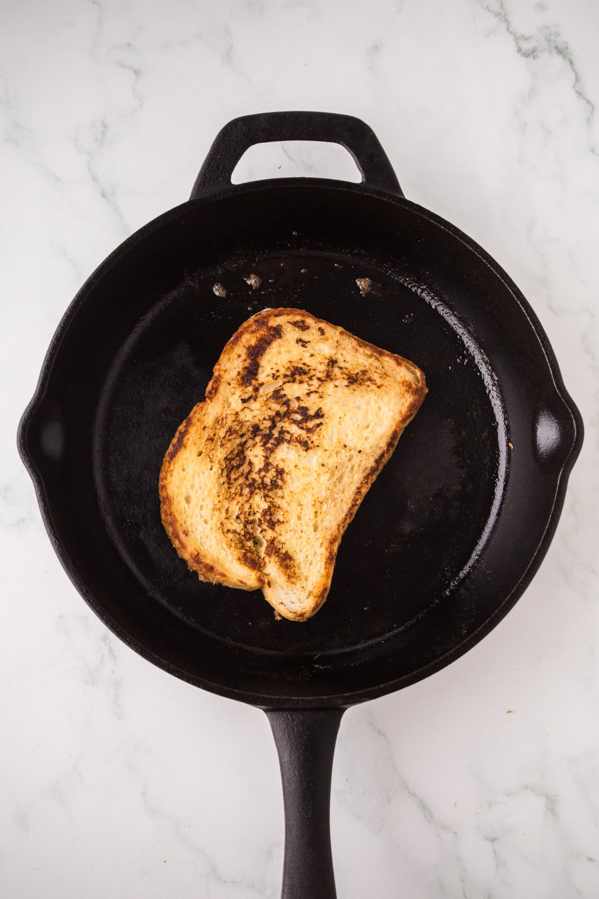 A single slice of sourdough French toast sits in the center of a black cast iron skillet on a white marble surface.