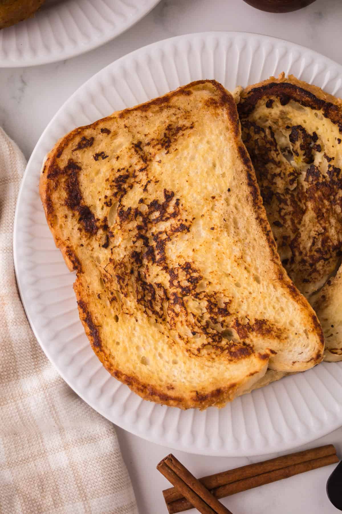 Two slices of golden-brown sourdough French toast on a white plate, with a beige napkin and cinnamon sticks nearby on a light surface.