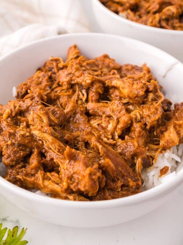 A white bowl filled with shredded chicken in a rich, reddish-brown sauce, inspired by a Thai chicken satay recipe, served over white rice. Another similar bowl and a sprig of parsley are visible nearby.