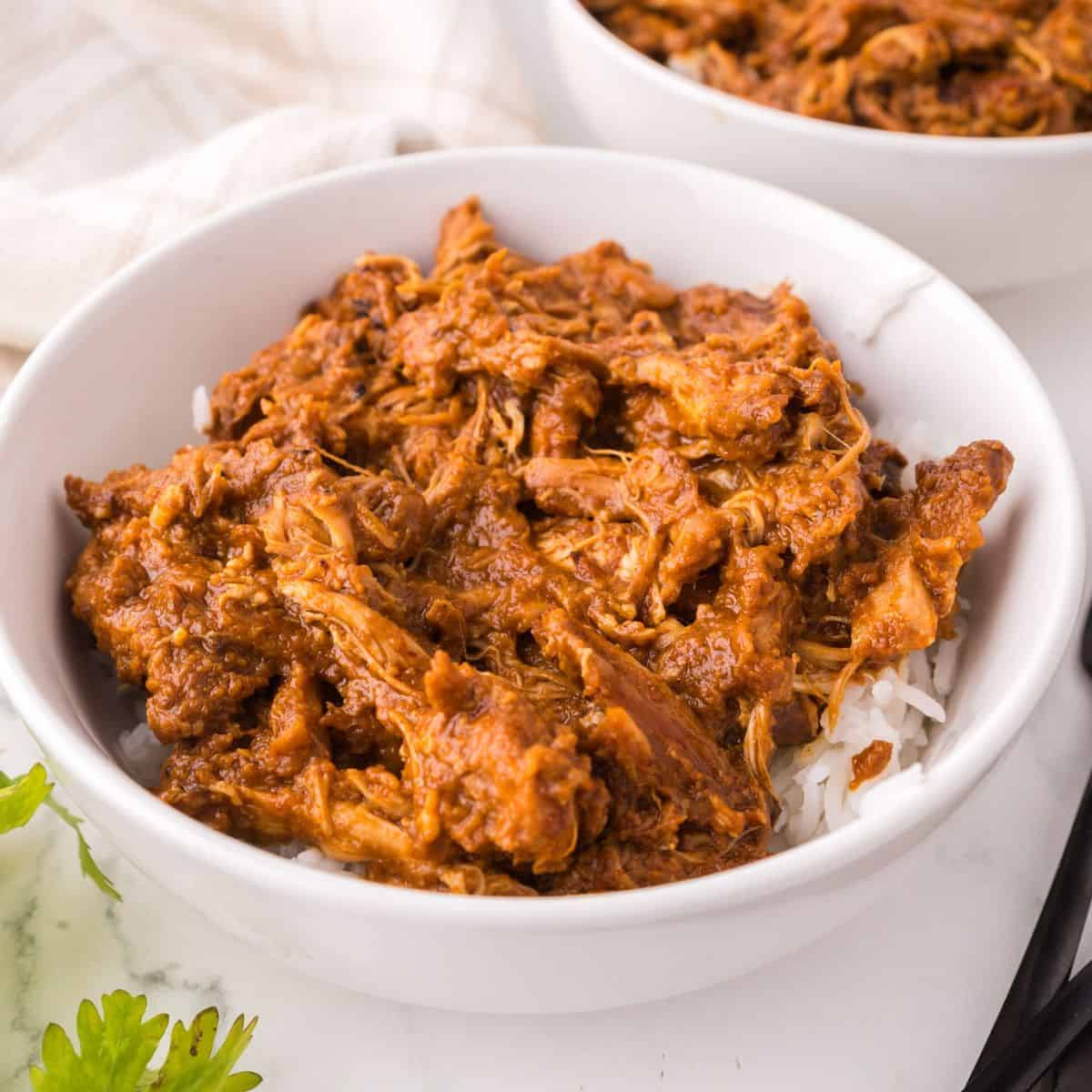A white bowl filled with shredded chicken in a rich, reddish-brown sauce, inspired by a Thai chicken satay recipe, served over white rice. Another similar bowl and a sprig of parsley are visible nearby.