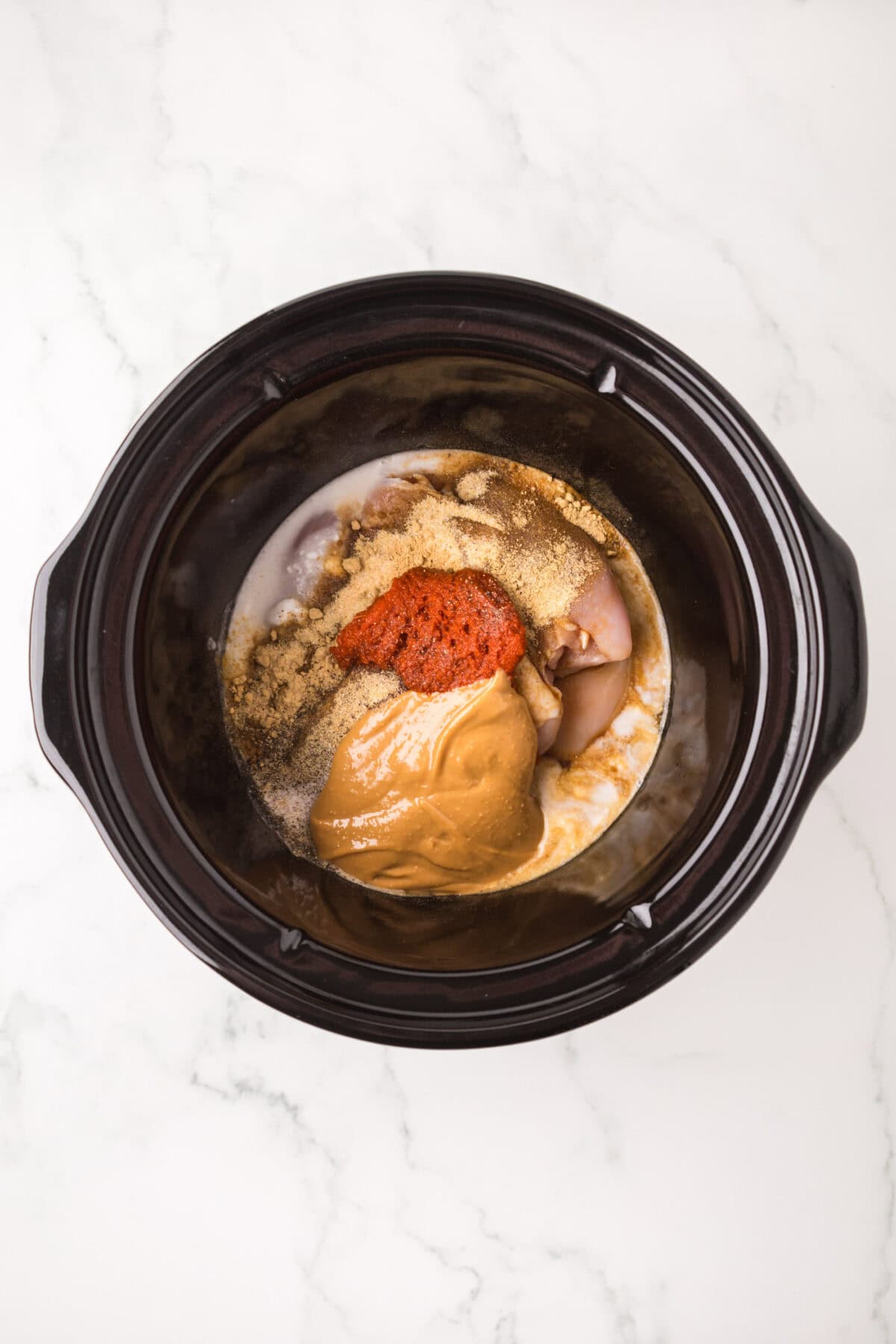 A slow cooker filled with raw chicken, peanut butter, spices, and seasonings for an easy Thai chicken satay recipe, ready to be mixed and cooked, placed on a white marble countertop.