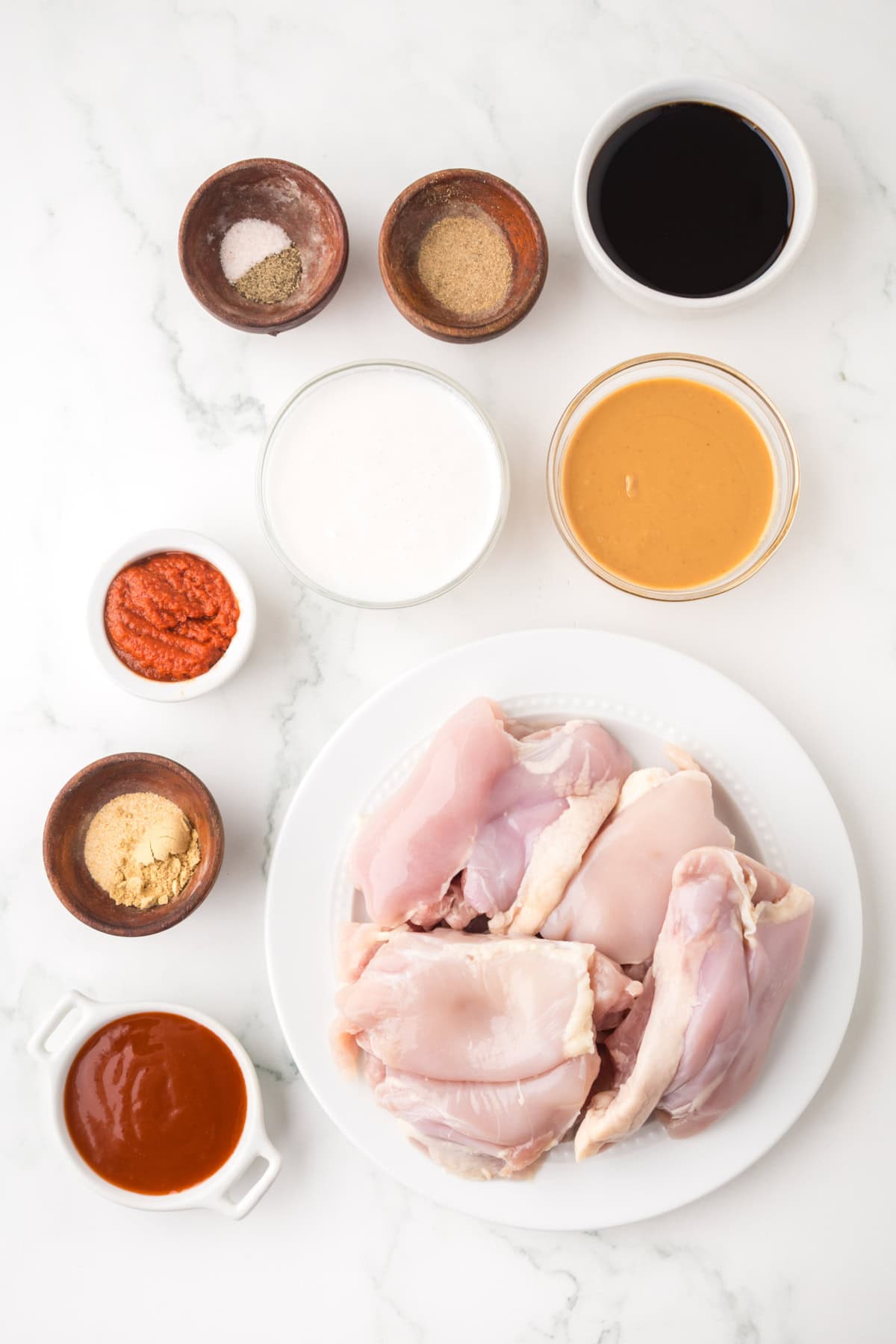 Raw chicken pieces on a white plate surrounded by small bowls of spices, sauces, seasoning, coconut milk, and peanut butter—classic ingredients for a flavorful Thai chicken satay recipe—arranged on a white marble surface.