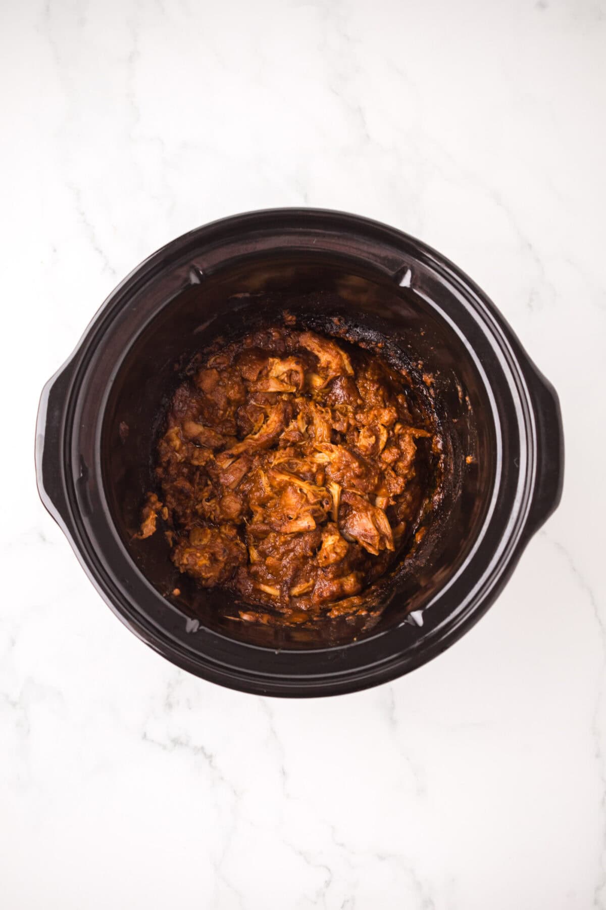 Shredded cooked meat in a rich, dark sauce reminiscent of a Thai chicken satay recipe, served inside a black slow cooker and placed on a white marble surface.