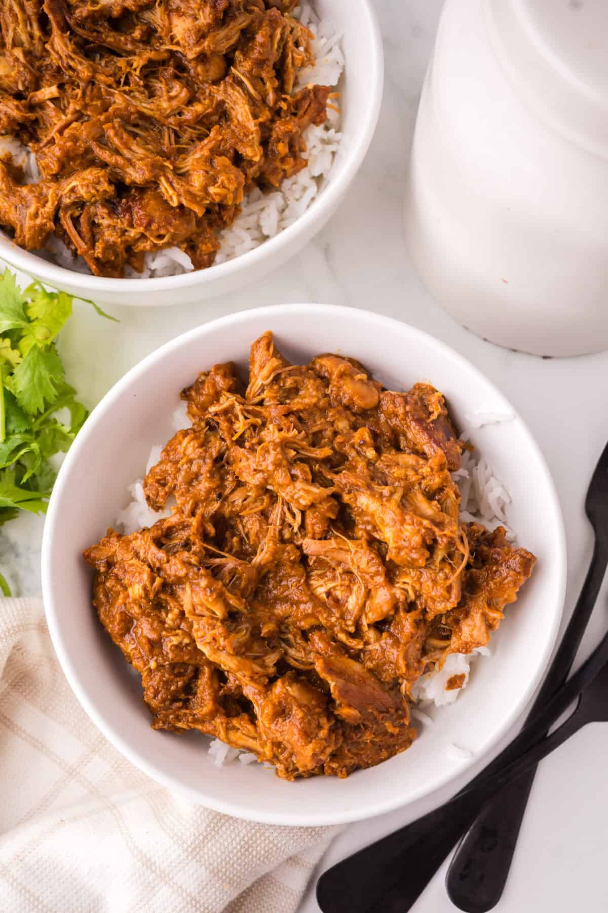 Two white bowls filled with shredded barbecue chicken, inspired by a Thai chicken satay recipe, served over white rice on a marble surface with black utensils, a beige cloth, and fresh cilantro nearby.