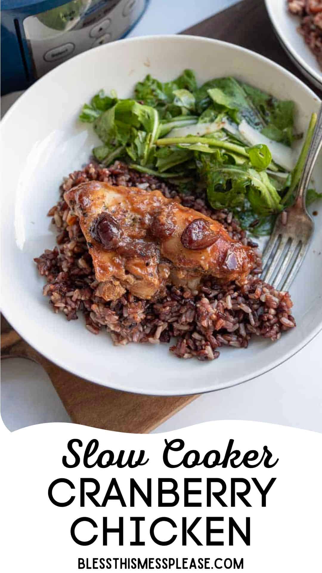 A plate of slow cooker cranberry chicken served on dark rice with a side of green salad, placed next to a fork. The cranberry chicken looks tender and saucy, with juicy cranberries adding extra flavor.