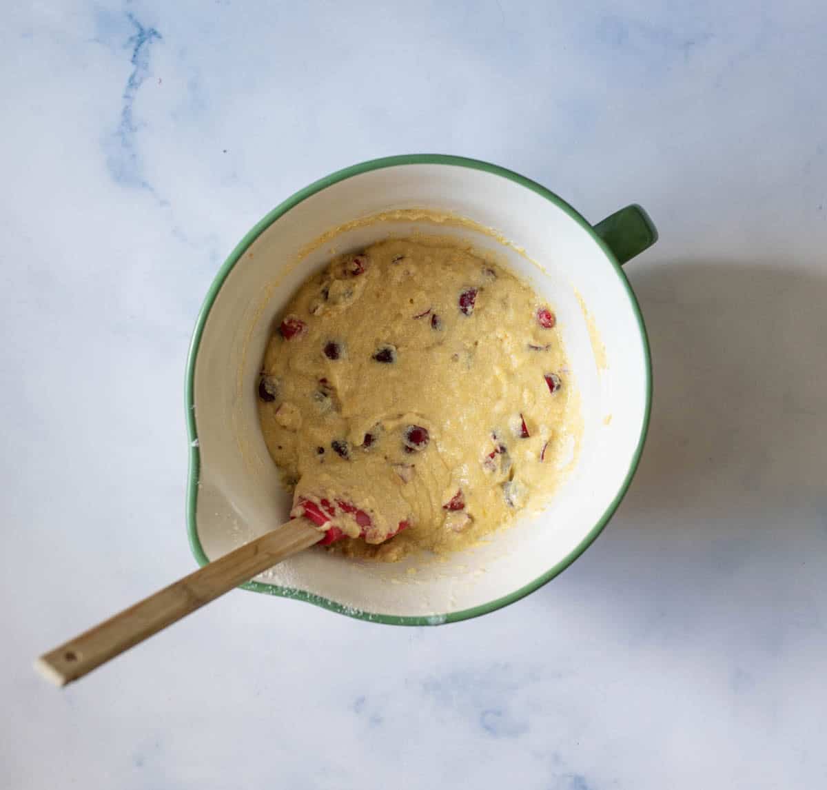 A white mixing bowl with a green rim contains thick batter studded with red cranberries, being stirred with a wooden spatula on a light, marble-like surface.