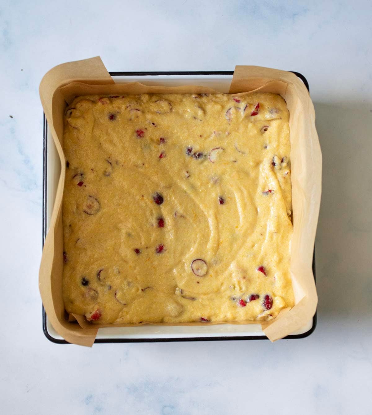 A square baking pan lined with parchment paper holds unbaked batter with visible pieces of fruit or nuts, set on a light marble surface.