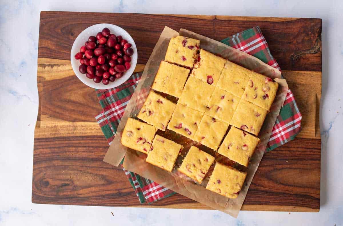 Squares of cranberry cornbread on parchment paper, partially cut, beside a small bowl of fresh cranberries on a wooden cutting board with a red and green plaid napkin.