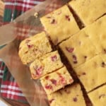 Squares of cranberry cornbread on parchment paper, with a few pieces stacked on the side. A red and green plaid cloth is partially visible underneath, and a small bowl of cranberries is at the top.