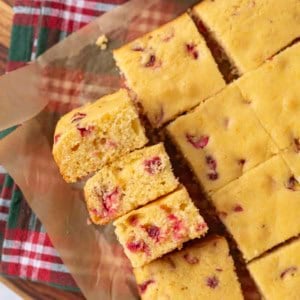 Squares of cranberry cornbread on parchment paper, with a few pieces stacked on the side. A red and green plaid cloth is partially visible underneath, and a small bowl of cranberries is at the top.