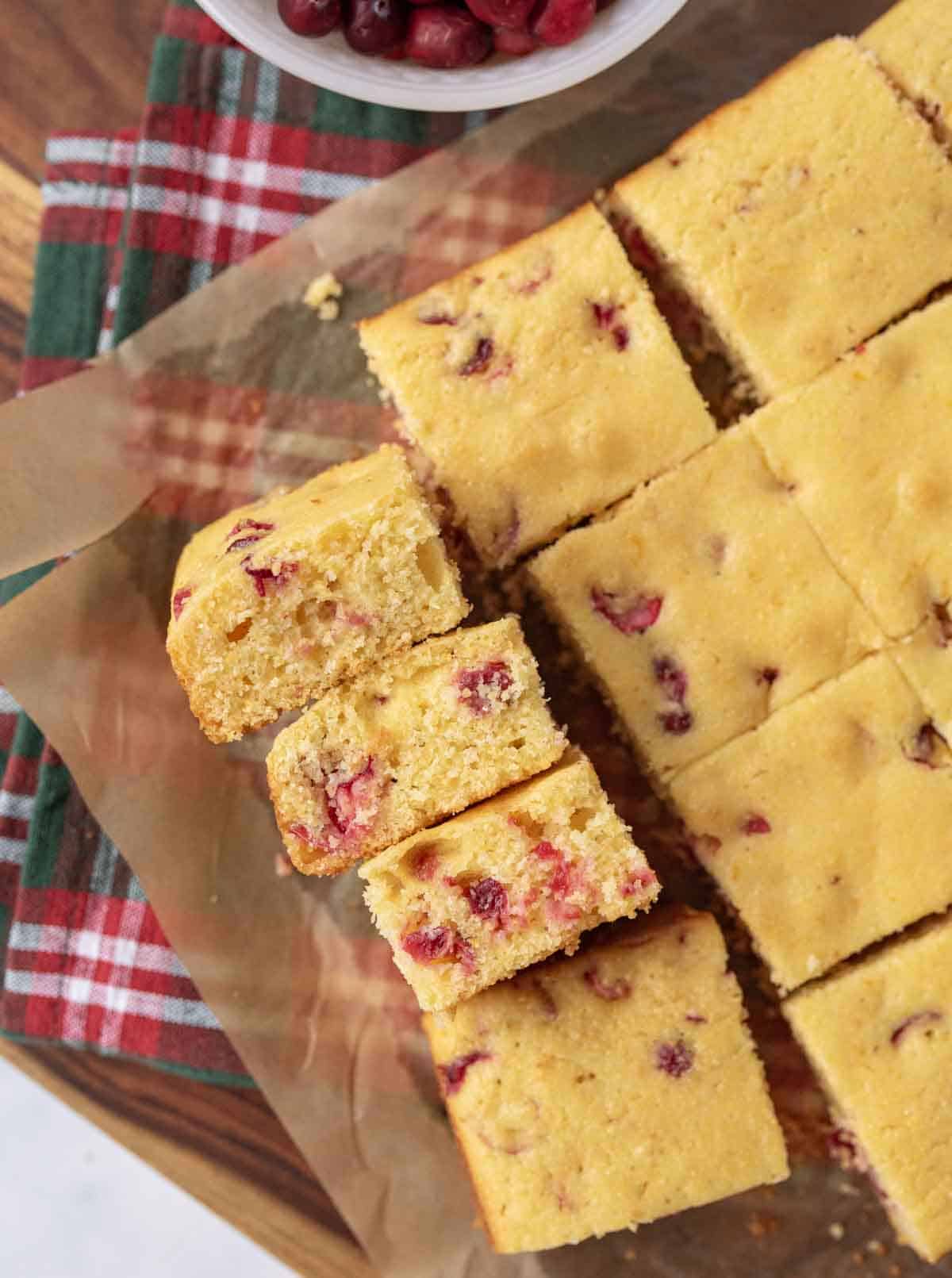 Squares of cranberry cornbread on parchment paper, with a few pieces stacked on the side. A red and green plaid cloth is partially visible underneath, and a small bowl of cranberries is at the top.