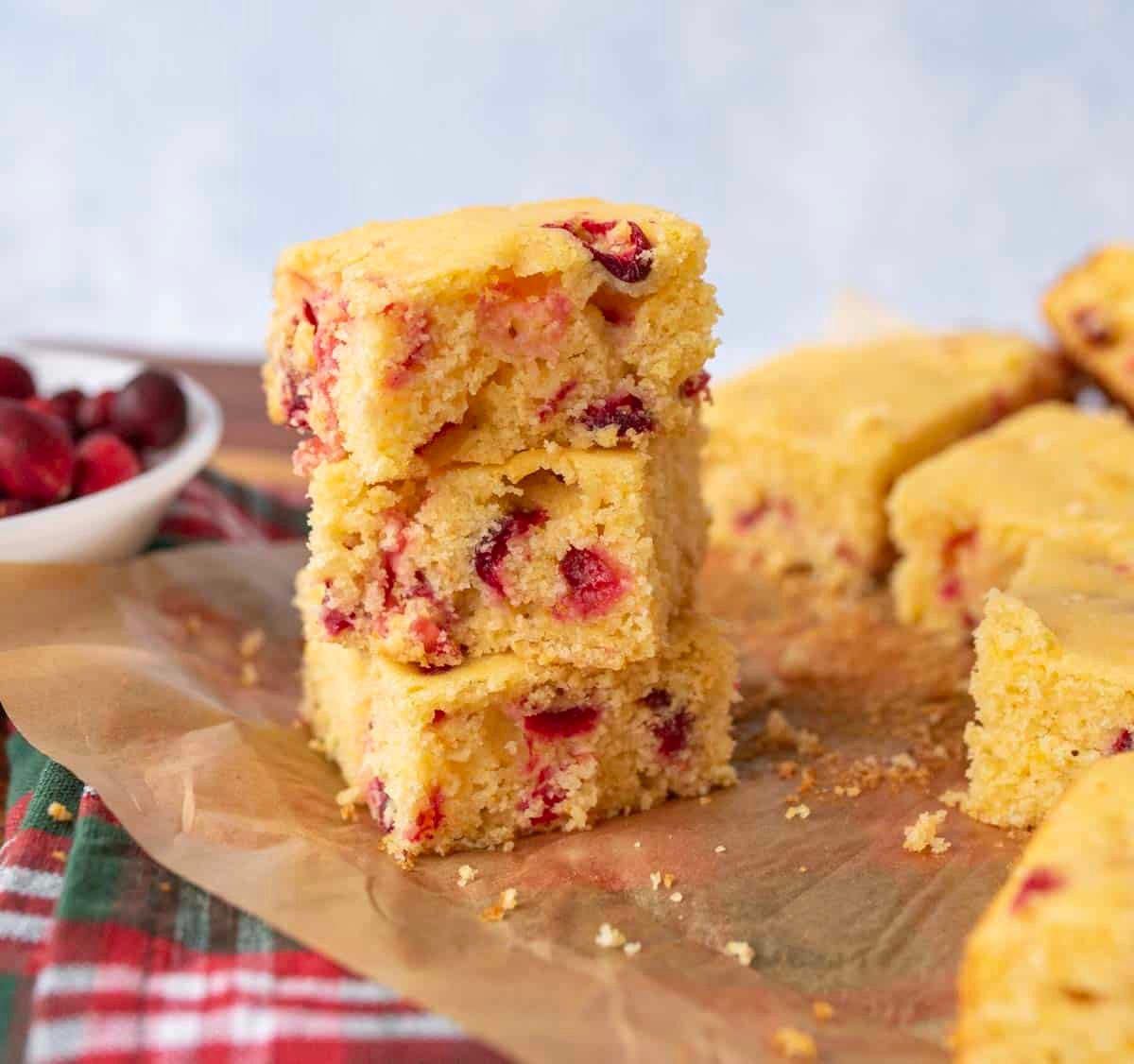 Three pieces of cranberry cornbread are stacked on parchment paper, with more cornbread squares and a bowl of cranberries in the background. The scene is set on a rustic surface with a plaid cloth.