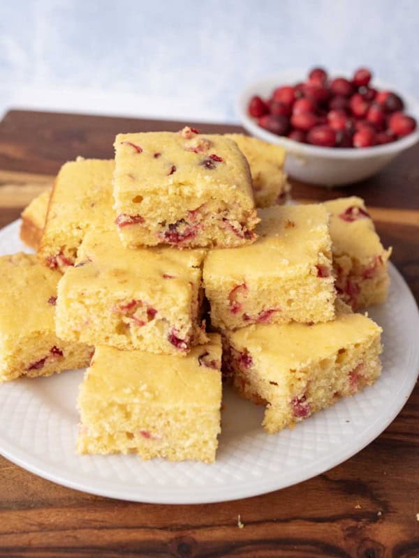 A white plate with a stack of square pieces of cranberry cornbread sits on a wooden surface. In the background, there is a small bowl filled with fresh cranberries.