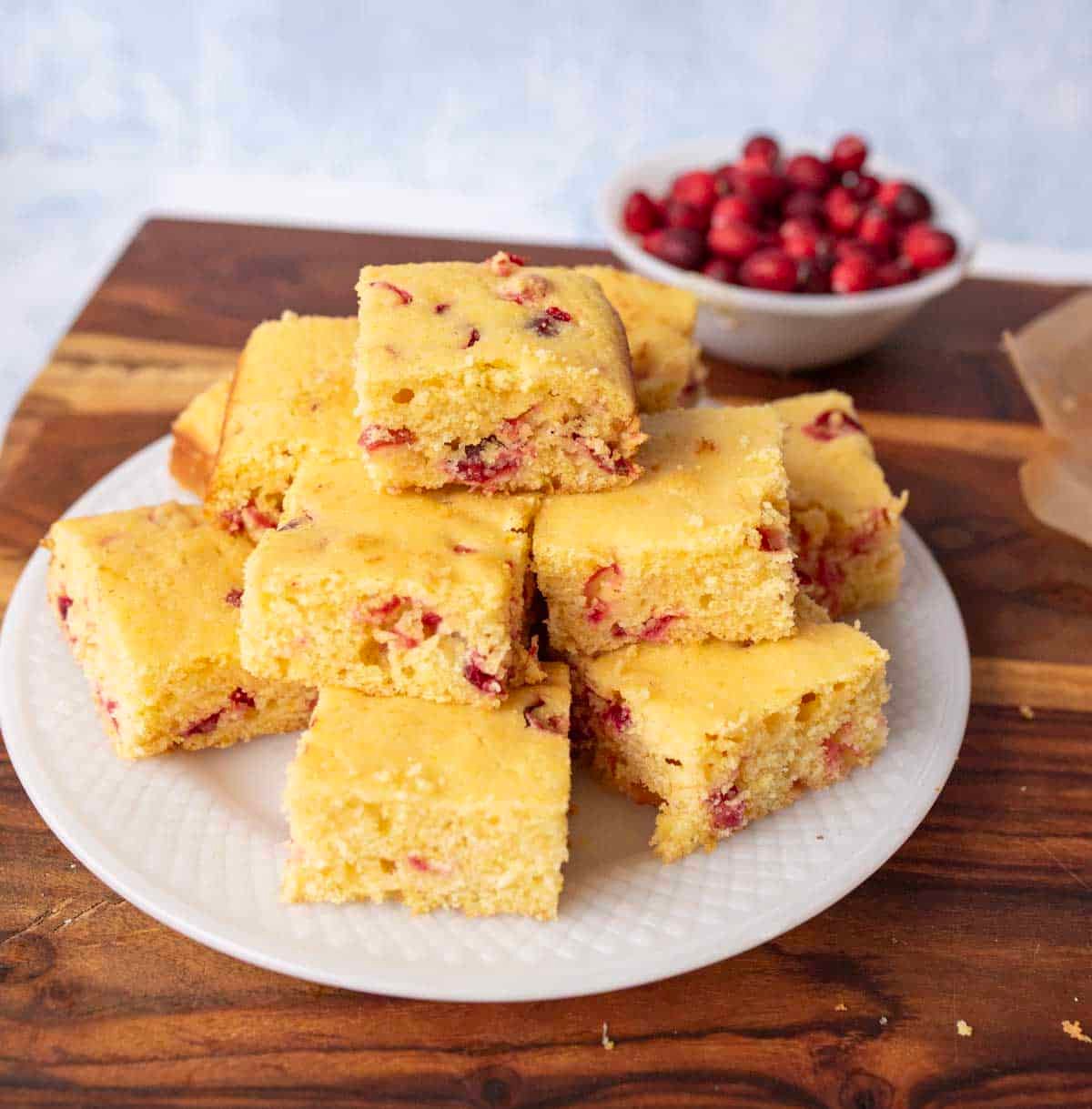 A white plate with a stack of square pieces of cranberry cornbread sits on a wooden surface. In the background, there is a small bowl filled with fresh cranberries.