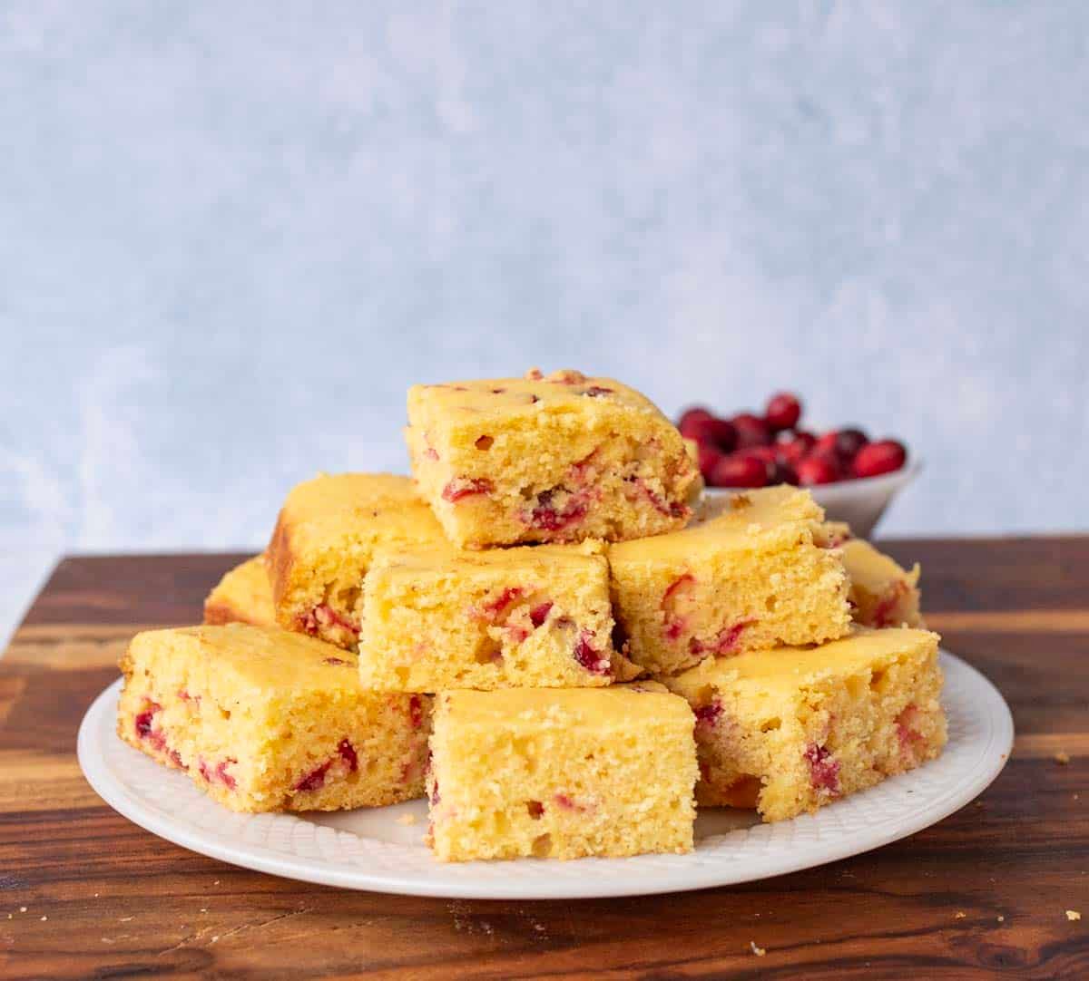 A plate of square pieces of yellow cranberry cornbread is stacked on a wooden surface, with a bowl of fresh cranberries in the background against a light blue wall.