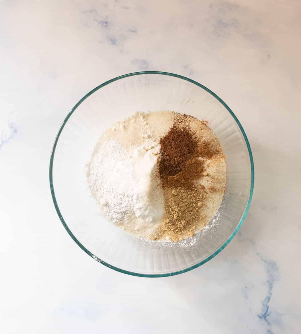 A glass mixing bowl containing dry baking ingredients, including white flour, sugar, and brown and tan spices, on a light marble surface.