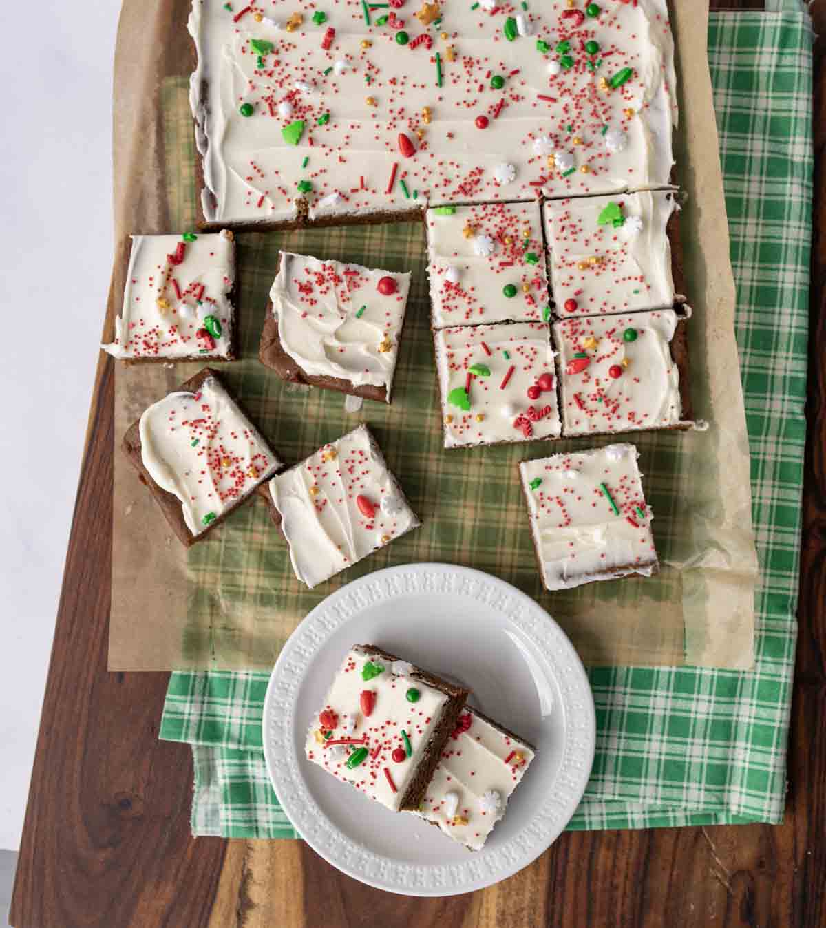 A tray of frosted brownies topped with colorful holiday sprinkles, cut into squares, sits on parchment and a green plaid cloth. Some brownies are arranged on a white plate and wooden surface.