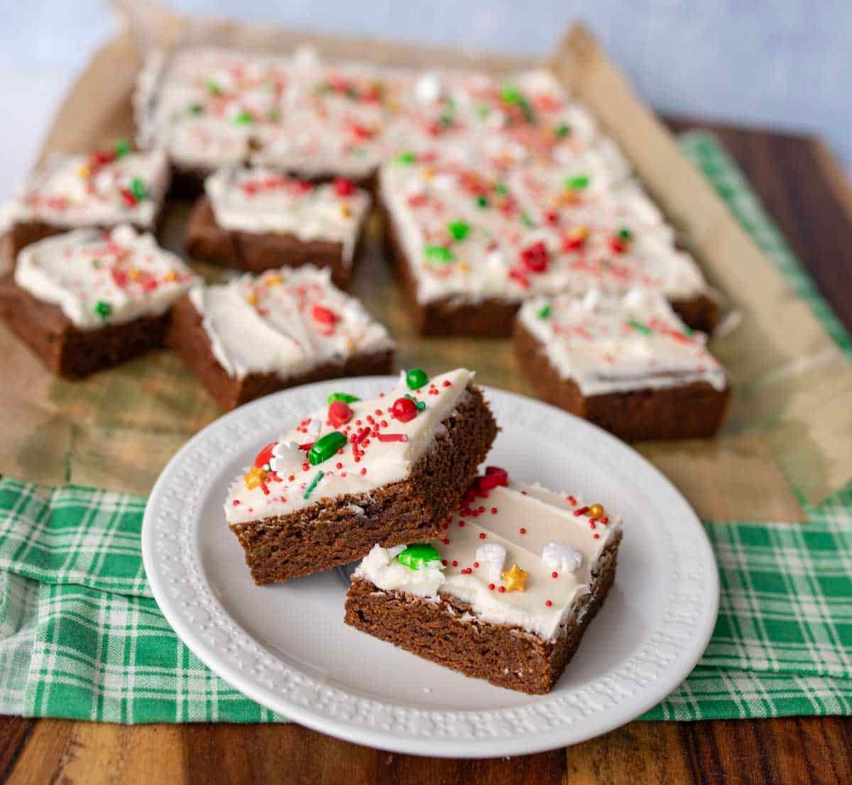 Two frosted gingerbread bars with colorful holiday sprinkles on a white plate, with more brownies on a parchment-lined tray in the background, all set on a green plaid cloth.