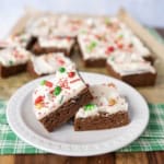 Frosted brownies topped with colorful red, green, and white holiday sprinkles are stacked on a white plate, with more brownies on parchment paper in the background. A green plaid cloth is underneath the plate.