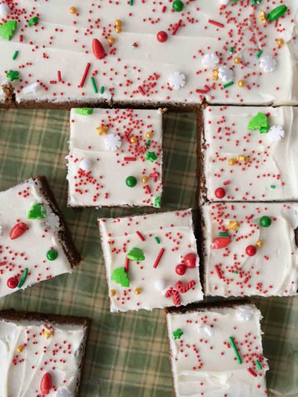 Frosted gingerbread bars cut into squares, topped with white icing and festive red, green, and gold holiday sprinkles, are arranged on a green plaid surface.